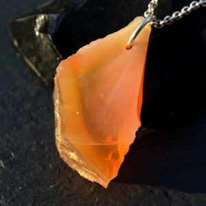 May include: A pendant necklace featuring a large, orange, and yellow, irregularly shaped, raw gemstone. The gemstone is attached to a silver chain.