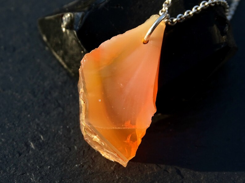 May include: A pendant necklace featuring a large, orange, and yellow, irregularly shaped, raw gemstone. The gemstone is attached to a silver chain.