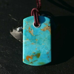 May include: Rectangular turquoise pendant with a maroon cord. The pendant has a polished surface with natural brown and orange veining. A silver-colored metal ring secures the cord to the pendant.