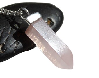 May include: A light pink crystal pendant necklace with a silver-colored metal cap and chain. The crystal is a pale pink color and has a faceted, rectangular shape. The necklace is displayed on a dark, textured surface.