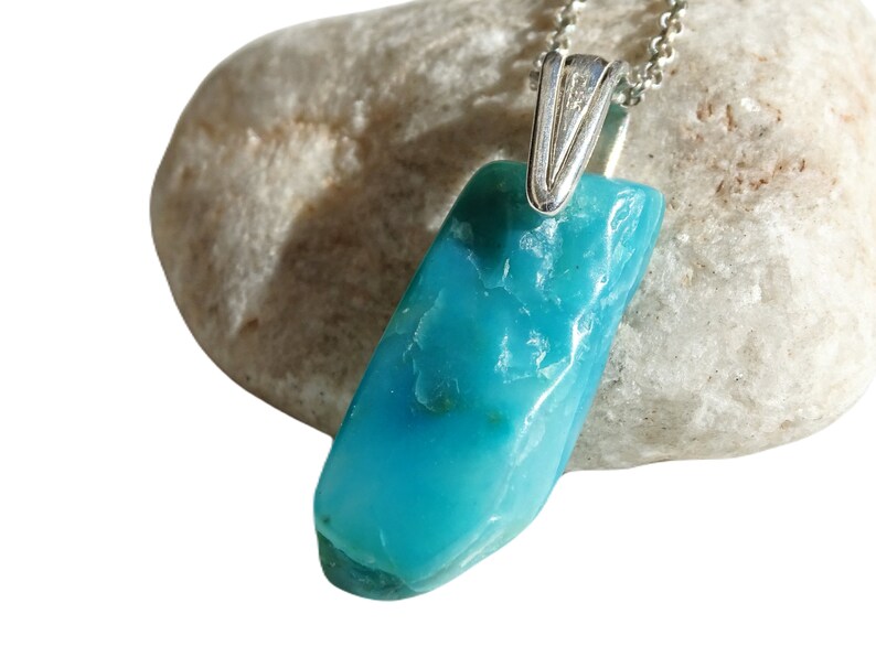May include: A turquoise blue gemstone pendant on a silver chain. The pendant is a rough, natural shape and is hanging from a silver bail.
