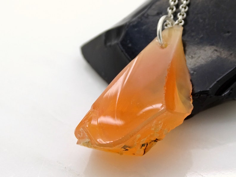 May include: A pendant necklace featuring a raw, orange carnelian stone with a silver chain. The carnelian is a natural, irregular shape with a smooth, polished surface.
