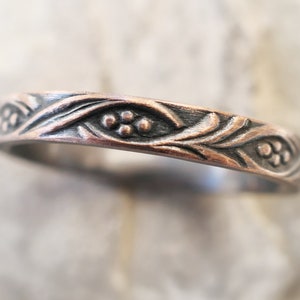 May include: A silver band ring with a floral design. The band is decorated with a repeating pattern of leaves and small flowers.