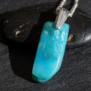 May include: A turquoise blue gemstone pendant on a silver chain. The pendant is a raw, irregular shape with a smooth, polished surface.