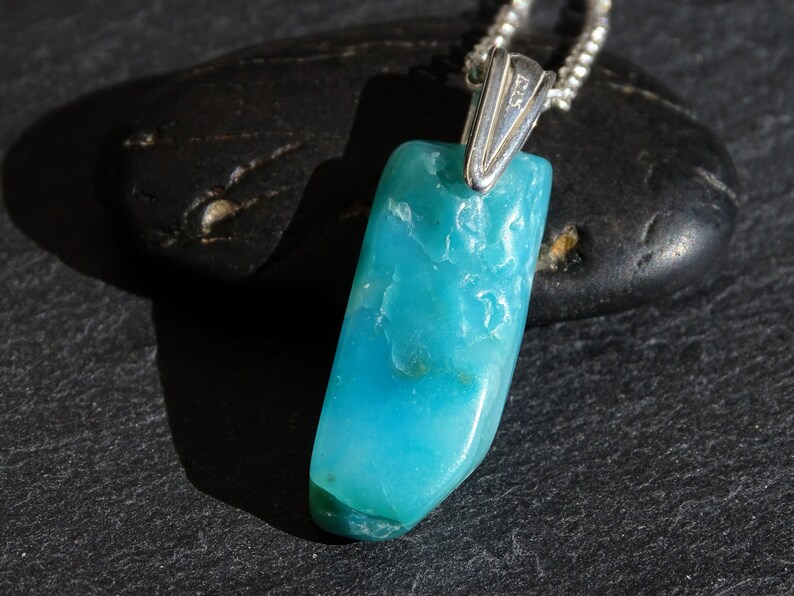 May include: A turquoise blue gemstone pendant on a silver chain. The pendant is a raw, irregular shape with a smooth, polished surface.