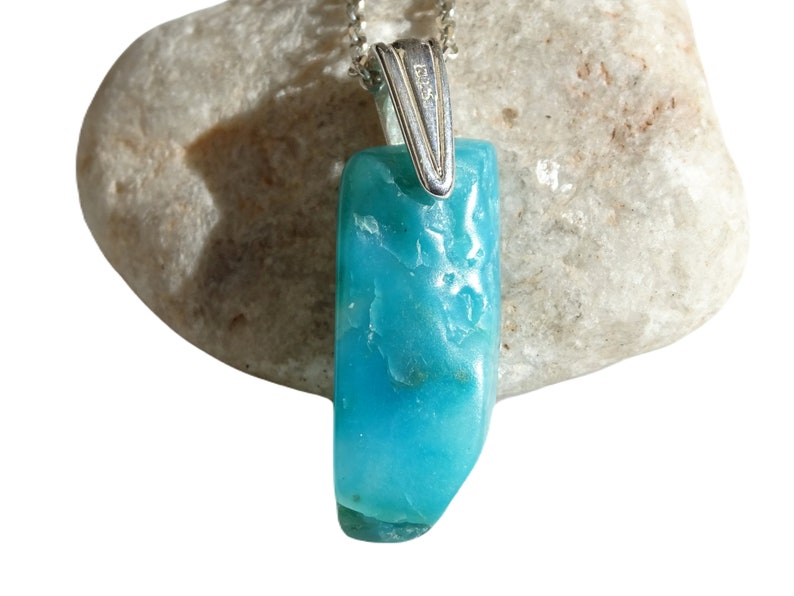 May include: A turquoise blue gemstone pendant with a silver bail. The pendant is on a white rock.