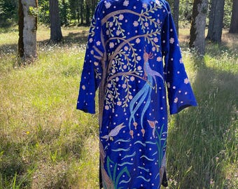 Pode incluir: Um longo robe azul royal com um design bordado de uma árvore, pássaros e elementos aquáticos. O robe é exibido em um manequim em um ambiente natural ao ar livre, mostrando seus detalhes intrincados e cores vibrantes.