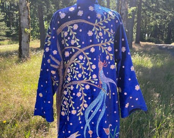 May include: A long, royal blue robe with a detailed embroidered design. The design features a tree with white flowers and a bird. The robe has long sleeves and a high collar. The background is a natural outdoor setting.