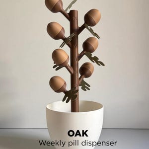 May include: A weekly pill dispenser shaped like a tree, with brown branches and acorn-shaped pill holders. The dispenser is in a white pot, with the word "OAK" and "Weekly pill dispenser" printed on the front.