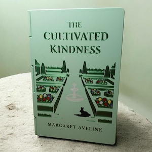 May include: A light green book titled "The Cultivated Kindness" by Margaret Aveline. The cover features a garden scene with a fountain, flower beds, and a silhouette of a rabbit. The book is resting on a textured surface.