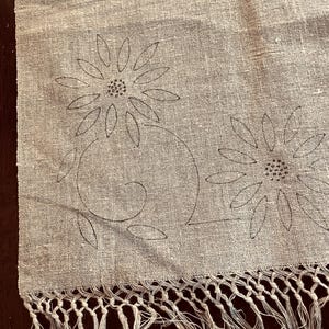 May include: A light beige woven fabric runner featuring two dark brown embroidered flower motifs with long petals and a central dotted circle, finished with a knotted fringe along the bottom edge of the textile.