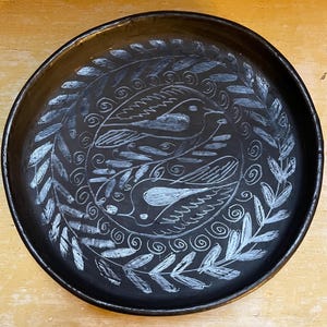 May include: A round, black ceramic plate with a white, hand-drawn design. The design features two birds surrounded by a wreath of leaves and branches. The plate has a slightly raised edge and is set against a light-colored wooden surface.