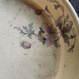 May include: A cream-colored ceramic bowl featuring a visible network of fine cracks across its surface. The interior is decorated with a floral motif in muted shades of purple, pink, and green, depicting a central flower with leaves.