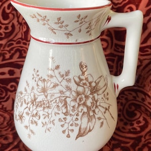 May include: White ceramic pitcher with a floral design in brown. The pitcher has a red rim and handle accents. The floral pattern includes flowers and leaves. The pitcher is a classic design for serving beverages.