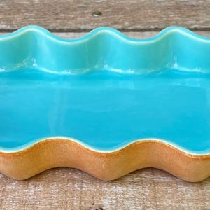 May include: A rectangular ceramic tray with a vibrant turquoise glazed interior and a wavy, scalloped rim. The exterior of the tray features a smooth, matte tan finish that contrasts with the glossy blue surface of the basin, creating a distinct look.