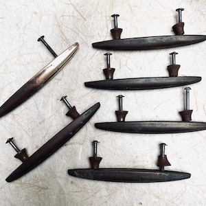 May include: A collection of six elongated drawer pulls with a dark, weathered metallic finish. Each handle features two mounting posts with attached screws. One pull is positioned diagonally, while the others are arranged in a vertical column.