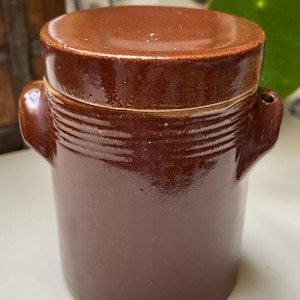 May include: A brown ceramic jar with a lid. The jar has two small handles and horizontal ridges near the top. The lid is a slightly lighter shade of brown. The jar is likely used for storage.