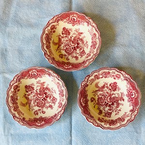 May include: Three round ceramic bowls featuring a cream-colored base with a deep red transferware pattern. The design displays intricate floral arrangements and stylized birds, finished with a scalloped edge and a decorative border pattern.