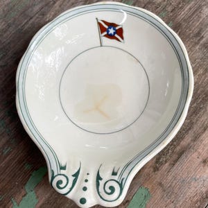 May include: White ceramic spoon rest with a blue and green trim. The spoon rest features a flag design with a red and white flag and a blue star. The handle has a decorative green design.