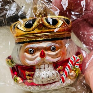 May include: A glass nutcracker ornament featuring a gold crown with black accents, a red jacket with gold glitter details, and a white beard. The figure holds a red and white striped candy cane and has a painted face with pink glitter eyebrows.