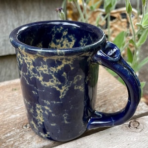 May include: A ceramic mug featuring a deep navy blue glaze with a mottled, sponge-like pattern in a pale cream color. The mug has a cylindrical body and a thick, curved handle attached to the side, showcasing a glossy, smooth finish throughout.