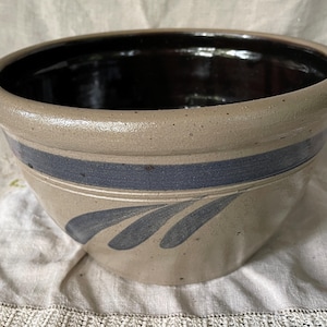 May include: A round stoneware mixing bowl with a speckled beige exterior and a dark brown glazed interior. The bowl features a wide, dark blue horizontal band around the upper portion and a hand-painted blue leaf-like design on the side.