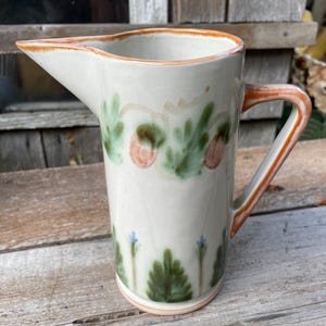 May include: A tall ceramic pitcher with a cream-colored glaze, featuring a hand-painted design of green leaves, orange acorn-like shapes, and small blue flowers. The pitcher has a prominent pouring spout and a curved handle with orange trim.