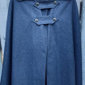 May include: A dark blue wool cape with a collar and three button closures. The cape has the initials "Y.H." embroidered on each side of the collar. The cape is hanging on a wooden fence.