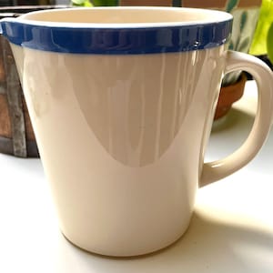 May include: A cream-colored ceramic pitcher with a smooth, glossy finish and a wide, dark blue band encircling the top rim. The pitcher features a curved handle on the right side and a small, integrated pouring spout on the left side of the rim.