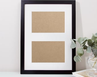 May include: A black double picture frame with a white mat and two rectangular openings. The frame is on a white surface with a vase of greenery and a stack of books in the background. The openings are filled with a light brown material.