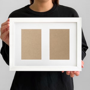 May include: A white picture frame with two openings, held by a person. The frame has a clean, modern design with a white border and a neutral-colored backing visible in the openings. The frame is designed to hold two photos.