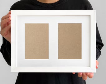 May include: A white picture frame with two openings, held by a person. The frame has a clean, modern design with a white border and a neutral-colored backing visible in the openings. The frame is designed to hold two photos.