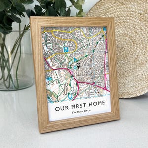May include: A framed map print in a light oak frame. The map shows a detailed street layout with a pin marking a location. The text "OUR FIRST HOME" and "The Start Of Us" are printed at the bottom of the map, perfect for a new home gift.