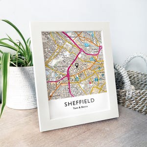 May include: A white framed print of a map of Sheffield, showing streets and landmarks. The map has pink and yellow detailing. The city name and a couple's names are printed below. A black location marker pinpoints a specific area on the map.