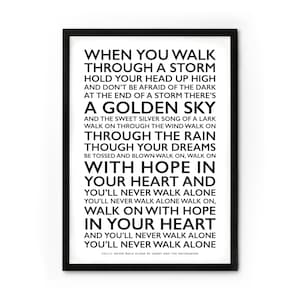 Könnte beinhalten: Ein gerahmter Druck mit schwarzem Rahmen zeigt den Text von "You'll Never Walk Alone" in schwarzer Schrift auf weißem Hintergrund. Der Text ist vertikal angeordnet und erzeugt einen auffälligen visuellen Effekt. Ein dekoratives Element.