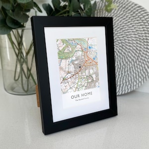 May include: A black framed print featuring a detailed map, with a pin marking a location. The print includes the text "OUR HOME" and "The Randell Family". The frame is propped up with a wooden stand.