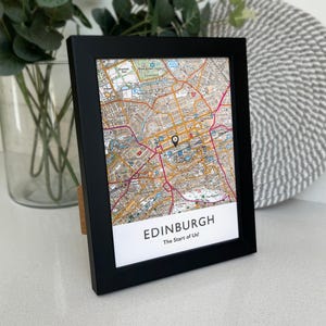 May include: A black framed print featuring a detailed map of Edinburgh, Scotland. The map shows streets and landmarks, with a black marker indicating a location. The text "EDINBURGH The Start of Us!" is printed at the bottom of the frame.