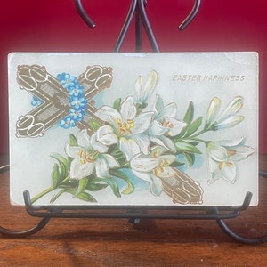Op de afbeelding: Vintage ansichtkaart voor Pasen met een gouden kruis versierd met blauwe vergeet-mij-nietjes en witte lelies met groene bladeren. De tekst "Easter Happiness" staat rechtsboven gedrukt.