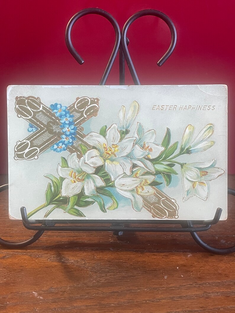 Op de afbeelding: Vintage ansichtkaart voor Pasen met een gouden kruis versierd met blauwe vergeet-mij-nietjes en witte lelies met groene bladeren. De tekst "Easter Happiness" staat rechtsboven gedrukt.