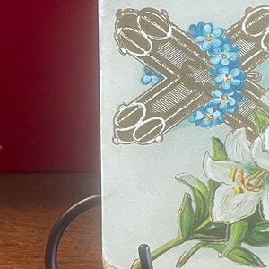 Op de afbeelding: Een vintage ansichtkaart met een gouden kruis met vergeet-me-nietjes en een witte lelie met groene bladeren.