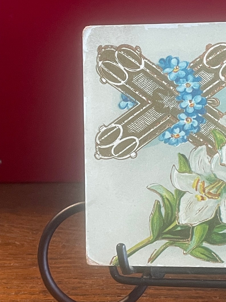 Op de afbeelding: Een vintage ansichtkaart met een gouden kruis met vergeet-me-nietjes en een witte lelie met groene bladeren.