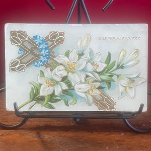 Op de afbeelding: Vintage ansichtkaart voor Pasen met een boeket witte lelies, een gouden kruis en blauwe vergeet-me-nietjes. De tekst "Easter Happiness" staat rechtsboven gedrukt.