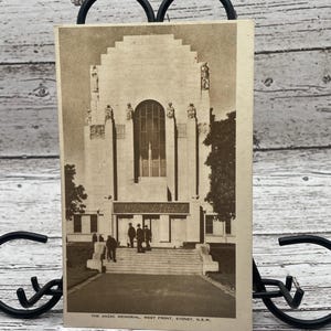 Può includere: Cartolina color seppia raffigurante l'Anzac Memorial a West Front, Sydney, N.S.W. L'immagine mostra la facciata del memoriale con una finestra ad arco e figure sui gradini. Gli alberi incorniciano l'edificio.