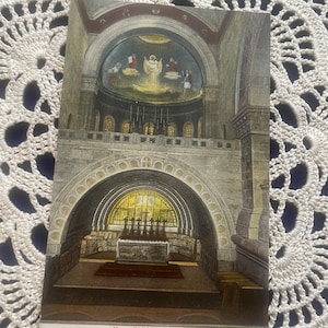 Puede incluir: Una postal vintage que representa el interior de la Basílica del Monte Tabor, en Israel. La imagen muestra un arco de piedra con una ventana de vidrieras y un altar cubierto con una alfombra roja. El techo presenta un fresco de Jesús ascendiendo al cielo.