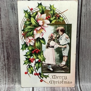 Può includere: Cartolina di Natale vintage con un'illustrazione a colori di un ragazzo e una ragazza, incorniciata da un bordo verde. La cartolina è decorata con agrifoglio, bacche rosse e fiori bianchi. Le parole "Merry Christmas" sono stampate in basso.