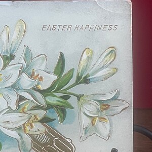 Op de afbeelding: Een vintage ansichtkaart met een boeket witte lelies en de tekst "Easter Happiness".