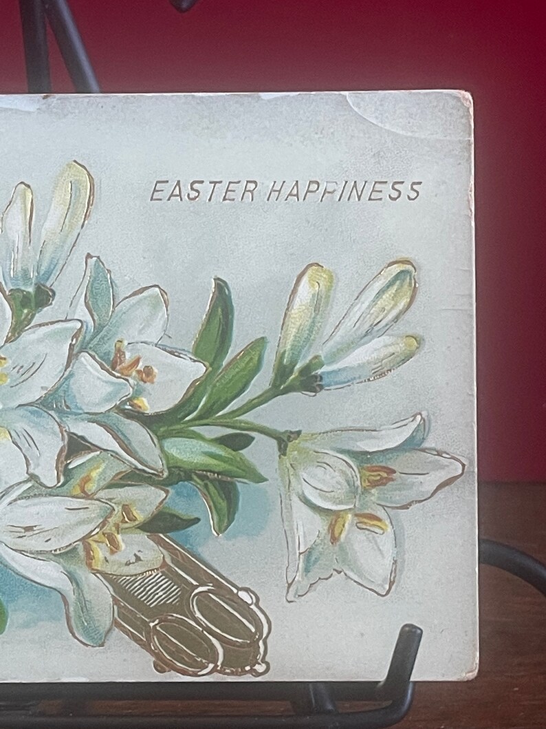 Op de afbeelding: Een vintage ansichtkaart met een boeket witte lelies en de tekst "Easter Happiness".