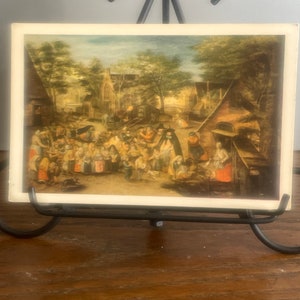 May include: A vintage postcard depicting a bustling village scene with people gathered in the street. The postcard features a colorful painting with a variety of buildings and trees in the background.