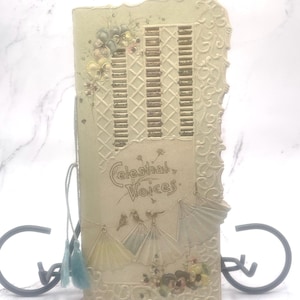 May include: A vintage, cream-colored card with a lace-like border and a gold glitter design. The card features the text "Celestial Voices" and a decorative design of birds and fans.