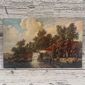 Könnte beinhalten: Eine Vintage-Postkarte mit einer Aquarellmalerei einer Wassermühle mit einem Wasserrad. Die Mühle ist von Bäumen und einem Bach umgeben. Der Himmel ist blau mit weißen Wolken. Der Text "THE OLD WATER WHEEL" ist unten auf der Postkarte gedruckt.